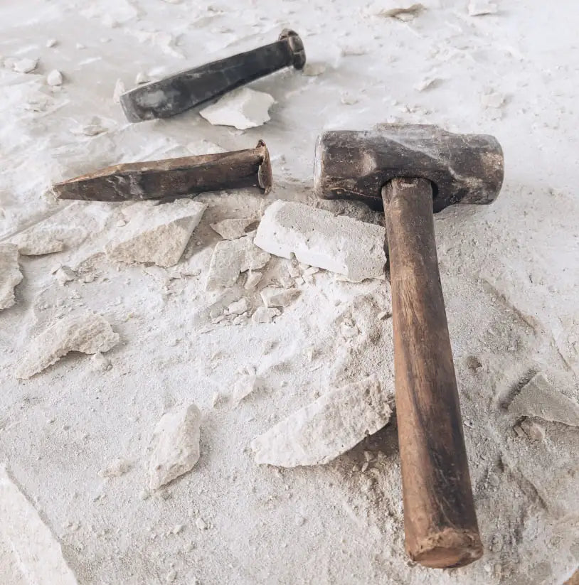 Hammer und Meißel liegen auf hellem Sandstein, umgeben von zerbrochenen Steinstücken, zeigen die Werkzeuge für die Bearbeitung von stonearts® Materialien.