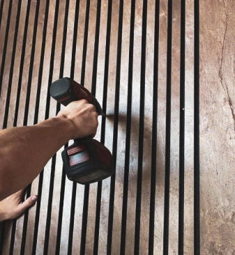 Person screws an Akurock acoustic panel from stonearts® to the wall, shows the simple installation of the acoustic panels with brown surface and black slats.